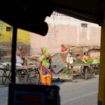 Experience the vibrant atmosphere of Agra's streets, where fresh produce and lively interactions bring the market to life. A vibrant street scene in Agra viewed from an auto rickshaw, showcasing a man at a fresh produce stall and a woman in a bright salwar kameez.