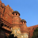 The majestic red sandstone Agra Fort, a testament to Mughal architecture. A low angle view of the Agra Fort's imposing red sandstone walls and towers against a clear blue sky, showcasing intricate carvings and lush greenery in the foreground.