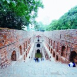 Exploring the Hidden Gems of Delhi: Agrasen Ki Baoli A group of people standing at the top of a stone staircase leading into an ancient stepwell, surrounded by lush greenery, perfect for touring Delhi.